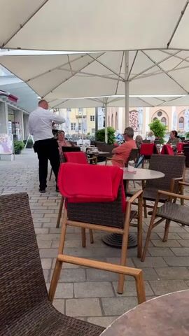 A man in a white shirt and black pants is walking away from the camera in an outdoor cafe setting. He appears to be a waiter, possibly carrying a tray, although it is obscured by his body. Several tables with chairs and red blankets are visible. In the background, other patrons are seated at tables, some talking and eating. The cafe is under a large white umbrella, providing shade. The ground is paved with cobblestones. The buildings in the background appear to be shops or residences, suggesting an urban or town environment. The lighting indicates it is daytime, likely overcast or shaded.