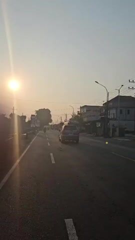 The video shows a road with traffic during sunset. The sun is low in the sky, casting a warm glow and lens flares. Several vehicles, including cars and trucks, are driving on the road. There are buildings and billboards visible on the sides of the road. The sky is clear and a light orange color. The overall scene depicts a typical sunset on a road with moderate traffic.