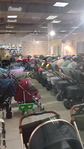 The video shows an indoor retail space, likely a baby store or a showroom specializing in baby strollers and prams. The primary focus is on a large display of numerous baby strollers arranged in neat rows throughout the store.

The strollers come in a variety of colors, including various shades of grey, black, olive green, beige, pink, and some with light blue patterned canopies. Many of the strollers have green star-shaped tags attached to their handles, possibly indicating a special offer, new arrival, or a featured product. Some of the strollers in the foreground feature stylish wooden or faux wood accents on their handles.

In the background, shelves are visible, displaying what appears to be more baby gear, possibly car seats, carriers, or additional stroller models. The store has a modern industrial feel with exposed ventilation ducts and fluorescent lighting fixtures on the ceiling. There's a prominent, bright light source on the right side of the frame, causing some glare in the video. The floor is light-colored and appears to be tiled.

The video slowly pans from left to right, providing a comprehensive view of the extensive selection of strollers available, highlighting the sheer volume and variety of options for customers. No people are visible in the store, suggesting it might be a quiet time or before opening hours.