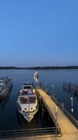 The video shows a wide shot of a body of water, likely a lake or a calm river, at dusk. The sky is a deep, clear blue, transitioning to darker shades towards the horizon. A line of trees is visible in the distance, forming a dark silhouette against the sky. In the foreground, a wooden pier extends into the water. There are several boats docked at the pier. The closest boat is a white and green passenger ferry with multiple decks and seating areas. It is docked stern-to the pier. To the left, another larger passenger boat is partially visible, also docked. Further out in the water, a smaller white boat with lights on is moving away from the pier, leaving a faint wake. The pier is illuminated by modern, downward-facing lights, casting a warm glow on the wooden planks. Some of the lights on the distant boat are also reflected in the water. The overall atmosphere is serene and peaceful, typical of the transition from day to night. There are no people visible in the video.