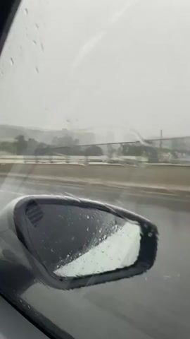 The video shows a view from inside a car driving on a highway in the rain. The windshield is wet with rain, distorting the view of the road and the surrounding environment. The side mirror on the right side of the car also shows rain on its surface. The highway has multiple lanes, and there is traffic visible in the distance. The weather appears overcast and hazy.