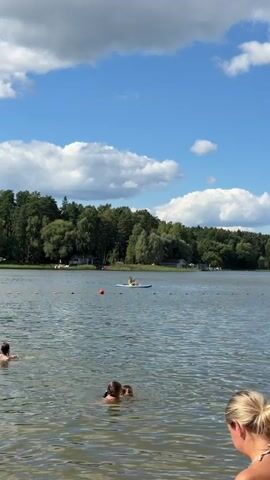 The video shows a scenic lake on a sunny day with scattered clouds. In the foreground, a woman with blonde hair pulled back is sitting on the shore, facing away from the camera. To her left, partially submerged in the water, are two women who appear to be swimming or wading. In the middle ground, several people are enjoying the water. One person is in a yellow kayak on the left side of the frame, and further to the right, a group of people are on a paddleboard. Other individuals are seen swimming or relaxing on inflatable rings in the water. In the background, a dense green forest lines the opposite shore. The lake is calm, with gentle ripples on the surface. Several buoys are visible, marking a designated swimming area. A white boat is docked on the far right of the lake.