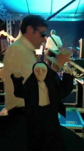 The video shows a nun figure standing on a blue platform in front of a band performing on stage. The nun figure is wearing a black habit and a silver cross necklace, and it is making the "rock on" hand gesture with both hands. Behind the nun, a band is playing. The band members are dressed in white shirts and dark pants. A man with sunglasses is playing the trumpet. A man is playing the drums, and two men are playing guitars. The stage is lit with blue and green lights. The video has a festive and lively atmosphere.