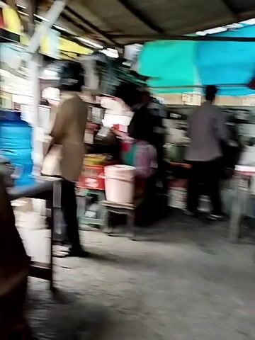 The video shows an indoor scene, possibly a food stall or a small eatery, with an open-air feel. The environment appears rustic and somewhat cluttered. In the foreground, a wooden table is visible. In the middle ground, a long table is set up with various bottles and containers of sauces and drinks. Several individuals are present. One person, wearing a grey t-shirt, is sitting at a table and drinking from a glass using a straw. Another person, wearing a blue t-shirt, is seated behind the first person. In the background, a person wearing a maroon shirt appears to be attending to something behind a counter. Another person, wearing a grey shirt and dark pants, is standing and facing away from the camera, near a cooking area with pots and pans. The ceiling is made of corrugated metal sheets, and there are blue and green tarps providing some cover. The floor is concrete. The overall impression is of a casual, functional, and possibly busy food establishment.