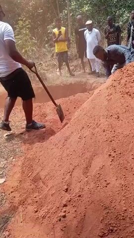 A group of approximately 10 people are gathered in an outdoor area that appears to be a wooded or partially cleared area. The ground is reddish-brown soil. Two men are actively digging a rectangular hole in the ground, presumably a grave. One man is using a shovel to dig and toss soil, while the other is using his hands to push soil out of the hole. The other people are standing around, watching the digging. The attire of the people varies, ranging from casual clothes like shorts and t-shirts to more formal wear. Some are looking at the hole, while others are looking around. There are trees and bushes visible in the background. The video appears to be taken during the daytime, as the scene is well-lit.
