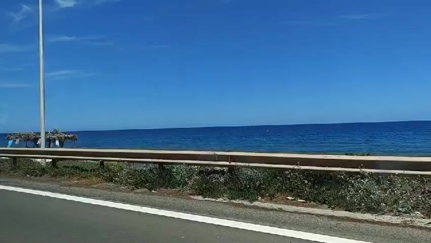 The video, filmed from a moving car, showcases a coastal road on a bright, sunny day. The clear blue sky and calm, deep blue sea create a serene backdrop. A road sign points to "Peroulia Beach" in Greek.  A simple, thatched-roof structure sits on the beach. The car's window is slightly dirty, with some water spots visible. A guardrail separates the road from the edge overlooking the beach. The vegetation along the roadside appears green and lush. The scene evokes a sense of travel and a sunny beach destination.