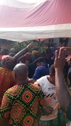 The video shows a group of people gathered under a red and white tent, appearing to be at an outdoor event. The attendees include both men and women, dressed in a variety of clothing styles, some traditional and some modern. One man in a black outfit, potentially security personnel, appears to be using a phone to record the gathering. The environment is outdoors, with houses visible in the background. Some people are seated, while others are standing.  The lighting seems to be daylight.