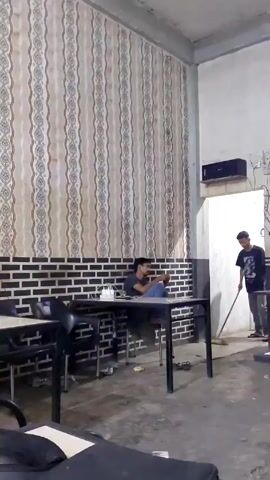 The video shows a room, possibly a restaurant, with two people. One man is seated at a table, looking at his phone. The other man is sweeping the floor near a doorway. The room has patterned wallpaper and a brick-like design on the lower part of the wall. There are several tables and chairs in the room. A speaker is mounted high on the wall near the doorway.