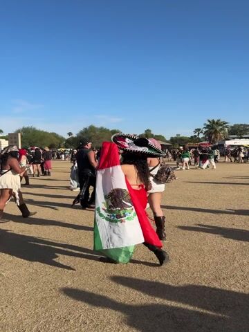 This video captures an outdoor festival scene during the daytime, under a clear blue sky. Numerous people are seen walking across a dusty, open field, suggesting a large gathering or event.

A prominent subject is a person, seen from behind, wearing a large sombrero and a Mexican flag draped over their shoulders like a cape. They walk among other festival-goers dressed in various casual and festive attire.

In another shot, a large, tall, cylindrical building with numerous rectangular window panels is visible. The windows appear to be made of colored glass or reflect light in a rainbow gradient, predominantly green and yellow. People are visible inside these windows, suggesting it's a viewing platform or an art installation. The word "PARADISE" is visible on one of the lower window panels of this structure. Palm trees are scattered in the background, indicative of a warm climate. The overall atmosphere is lively and festive.