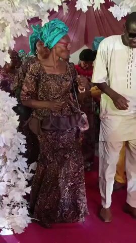 A short video shows a group of adults dancing under a wedding arch. A man in a white suit and sunglasses dances to the right, and a woman in a brown, sequined dress and a green headwrap dances to the left. Other women, also in green headwraps and similar dresses, are visible behind them. The arch is decorated with white, leaf-shaped decorations. The scene appears to be a wedding celebration.