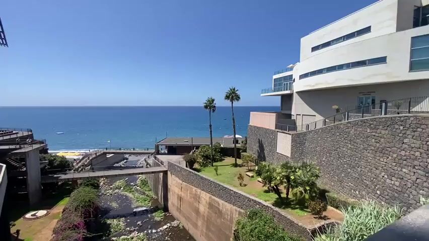 The video shows a sunny day overlooking the ocean. In the foreground, there is a concrete channel with a small stream flowing through it. There are buildings with modern architecture on both sides of the channel. Palm trees are visible in the background. The ocean is calm and blue, with a beach and some boats visible. The sky is clear and bright blue.