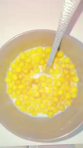 A high-angle, static shot shows a bowl of cereal. The bowl is dark grey and contains yellow, round cereal pieces submerged in milk. A silver spoon with a textured handle is inside the bowl. There is a sprinkling of brown powder, possibly cinnamon, on top of the cereal. The bowl sits on a white surface with a hint of pink in the top left corner.