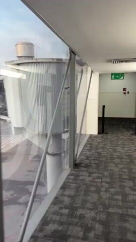 The video begins inside an airport jet bridge. On the left, through a large glass window, an airplane is visible parked at the gate. Several ground crew members are working around the plane. The jet bridge has a carpeted floor and is enclosed by glass panels on the left, with a metal railing running along the glass. The camera pans to the right, showing the interior of the jet bridge. The floor is carpeted with a grey and black patterned carpet. Further down the jet bridge, there are two black stanchions with red tops. An emergency exit sign with a green running figure is visible on the wall ahead. The video continues to pan and appears to show the end of the jet bridge with a doorway ahead.