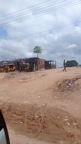 The video depicts a scene from what appears to be a market area or a roadside settlement in a developing country. The ground is dusty and unpaved. Several yellow three-wheeled vehicles (auto rickshaws or "keke napeps") are parked along the side of the road. Makeshift stalls and structures are lined up behind them, suggesting a market or informal trading area. The sky is cloudy. A large blue and white semi-truck passes by on the road. A red pickup truck and stacks of used tires are also visible.