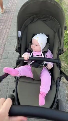 A baby is sitting in a dark green stroller, looking to the left. The baby is wearing a white hat with yellow stars and pink ribbed pants and socks. The stroller has a black bar in the front that the baby is holding onto. The stroller is on a paved path with grass on the right side. A person's hands are holding the stroller handles, with the right hand closer to the camera. The video is filmed from the perspective of the person pushing the stroller.