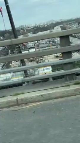 The video shows a view from a moving vehicle on a bridge. The bridge railing is visible in the foreground. Beyond the railing, there's an industrial area with warehouses, containers, and construction equipment. In the distance, there is a city skyline under an overcast sky.