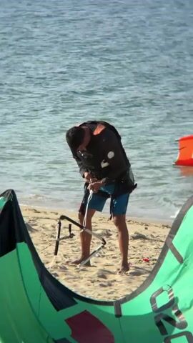 A man in a black jacket and blue shorts, wearing a life vest, stands on a sandy beach near the water, adjusting a kiteboarding bar. In the background, another person in the water with an orange wing and a board, and a green and black kite lie on the sand in the foreground. The water is calm with gentle waves.