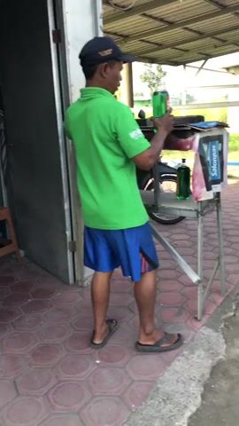 A man in a green shirt and blue shorts is holding a green soda can and a red balloon, seemingly about to pop the balloon with the can. He is standing on a tiled surface next to a building.