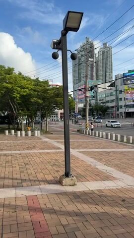 A short video clip captures a street scene in what appears to be a city. The foreground features a paved walkway with red and beige brick-like tiles laid in a pattern, with concrete borders. A tall, dark gray lamp post stands prominently in the center of the foreground, its base resting on a concrete slab. The lamp post has a rectangular light fixture at the top and two smaller spherical objects attached to its pole, one above the other. Behind the lamp post, a street can be seen with several cars driving on it. The street is bordered by white cylindrical bollards. In the middle ground, there is a crosswalk and traffic lights. A few pedestrians are visible on the sidewalk and crossing the street. A person is sitting on a low wall near the street. In the background, there are several multi-story buildings, including a tall, modern skyscraper with a distinctive architectural design. The sky is blue with scattered white clouds. Power lines are visible crisscrossing the sky. Some of the buildings have large advertisements or signage displayed on them. The overall impression is a typical urban environment on a clear day.