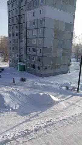 The video pans across a snowy, urban landscape featuring two Soviet-era apartment buildings, several parked cars covered in snow, and a low brick building with boarded-up windows. The ground is entirely covered in snow, with tire tracks visible on the road. A figure walks in front of one of the apartment buildings, carrying what appears to be a bundle of sticks or branches. The sky is clear and bright.