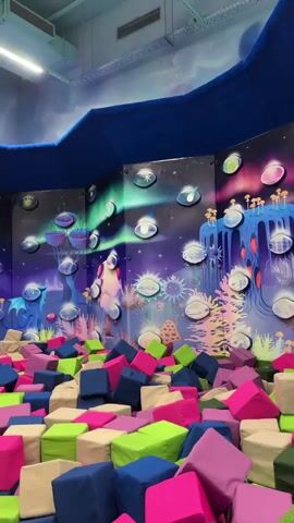 This video captures a vibrant indoor playground, likely a children's entertainment center. The walls are adorned with fantastical murals depicting outer space scenes, complete with planets, astronauts, and whimsical creatures, suggesting a playful and imaginative atmosphere. The primary objects in the foreground are a large foam pit filled with an assortment of brightly colored foam cubes in shades of pink, green, and blue, creating a soft and inviting play area.

In the background, a trampoline area with a blue and green padded surface is visible, framed by safety netting. A few individuals, appearing to be children, are engaged in play on the trampolines. Their movements are dynamic and energetic, indicating active participation and enjoyment. One shot shows a girl in a white shirt and jeans on the trampoline, mid-motion, possibly jumping or preparing to jump. Another sequence features a blurry shot of children interacting with the foam pit and trampolines.

The setting is indoors, illuminated by artificial lighting, suggesting it could be any time of day. There are no cues about weather conditions. The overall mood conveyed is one of fun, excitement, and active recreation, typical of a children's play facility. While the specific location is identified as Sovkhoza imeni Lenina, Russia, there is no visible text in the video itself to confirm this or provide any other information.