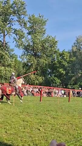 The video captures a jousting tournament. Two knights on horseback, clad in full armor and carrying lances, charge towards each other in a grassy field. Spectators are visible in the background, seated behind a wooden fence. The trees provide a natural backdrop to the event. The knights are focused on the competition, their lances aimed at their opponents. The atmosphere appears festive, with the sun shining brightly on the scene.