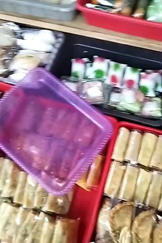 A hand is placing a small, clear plastic bag containing a brown liquid and what appears to be spices into a purple plastic tray. The tray is situated in a market stall filled with various food items. To the left of the tray are numerous individually wrapped food items, some are long and cylindrical, possibly spring rolls or similar snacks, and others are more square or rectangular. To the right of the tray, there are also rows of wrapped food items, and behind the purple tray, there are more food items displayed, including what look like colorful layered cakes or pastries. The overall environment appears to be an outdoor or semi-enclosed market stall with a focus on pre-packaged food.