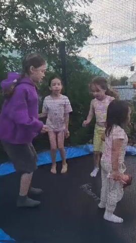 Four young girls are jumping on a trampoline outdoors. The trampoline is round with a blue padded edge and a black jumping surface, surrounded by a safety net. In the foreground, a girl in a purple hooded sweatshirt and dark shorts is facing away from the camera, her hair in a ponytail. To her left, another girl with dark hair in a ponytail is wearing a white t-shirt and pink shorts. She is mid-jump, with her legs bent and arms raised. To the right of the first girl, a girl in a yellow crop top and yellow shorts is also jumping, with her arms outstretched. In the far right foreground, a girl with dark hair is crouched on the trampoline, facing towards the center. The background shows green trees, bushes, and a glimpse of a house and fencing. The sky is visible, with some clouds. The lighting suggests it is daytime, possibly late afternoon or early evening. The girls appear to be interacting and playing together on the trampoline.