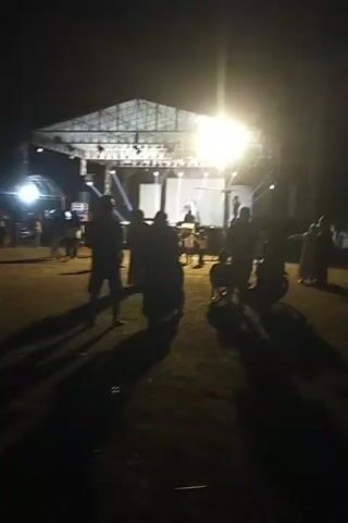 The video shows an outdoor event at night. There is a stage set up with bright lights, and a crowd of people are gathered in front of it. Some people are standing, while others appear to be sitting. There are also several motorcycles parked in the foreground. The overall impression is of a lively gathering, possibly a concert or festival. The lighting suggests it is dark outside, and the stage lights are the primary source of illumination for the event area. Shadows are cast on the ground from the people and objects, indicating the direction of the light sources. The ground appears to be a mix of grass and dirt.