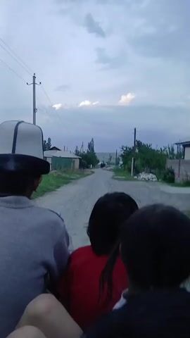 This video captures a journey on a dirt road in Kotormo, Kyrgyzstan. The perspective is from the back of a moving vehicle, likely a cart or truck, with several people inside. In the foreground, we see the backs of several individuals, including a man wearing a white hat with black trim, a person in a red shirt, and others with dark hair. The road ahead stretches into the distance, lined with modest houses, fences, and trees. Utility poles with wires are visible along the roadside, indicating a rural or semi-rural setting. The sky is overcast with streaks of light, suggesting it is either early morning or late afternoon. The overall atmosphere appears calm and contemplative, with the focus on the passage through the village. The activity is simply travel along this path. No visible text is present in the footage.