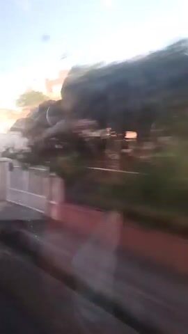 The video shows a blurred view of houses and trees passing by, suggesting it was filmed from a moving vehicle. The sky is visible and appears to be a light blue with some clouds. The houses have tiled roofs and are white or light-colored. There are fences and some vegetation along the side of the road. The motion blur indicates a relatively fast speed. There are no clear people or distinct actions visible due to the blur.