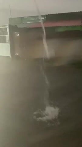 A person is standing in the water in front of a building and a fence. The person is wearing a dark shirt and shorts. Water is pouring down from above, hitting the person and creating a splash. The surroundings are dark and misty.