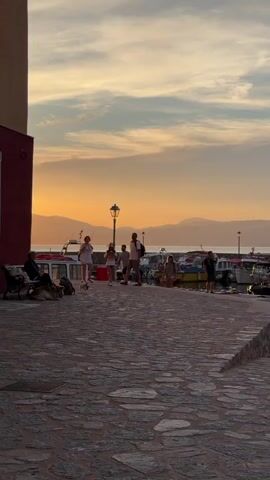 A group of people are walking along a paved walkway in front of a harbor at sunset. The sky is orange. There is a person sitting on a bench on the left side of the frame, with a dog lying at their feet. The group includes a man, a woman, and two girls. They are all dressed in casual clothing. A small white dog walks in front of the group. Behind them, boats are docked in the harbor. Mountains are visible in the distance. There is a lamppost in the center of the frame.