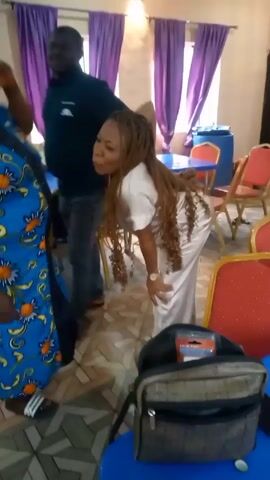 The video shows a room with a tiled floor, purple curtains, and blue and red chairs. Several people are moving around, some are laughing and talking. A woman in a blue dress with orange flowers is standing and talking to another woman in a white dress. A man in a dark shirt and grey pants walks past. There is a table with food on it in the foreground. The room appears to be set up for an event.