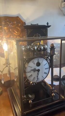 The video shows a close-up view of a complex clock mechanism with a glass enclosure. The clock has a white face with Roman numerals and ornate hands. Above the clock face, a series of metal balls are arranged on a tiered platform. Below the clock face, a pendulum or similar oscillating mechanism is visible, along with a small circular object labeled "LIC". The clock is set against a background of antique furniture and decorative items, including a gilded mirror or frame and a dark, ornate cabinet. The lighting in the scene is warm, with a bright light source visible to the left, casting a glow on the clock and its surroundings. The camera angle is static, focusing on the clock's intricate details and its operation. The overall impression is one of a vintage, mechanical timepiece displayed in a richly decorated setting.
