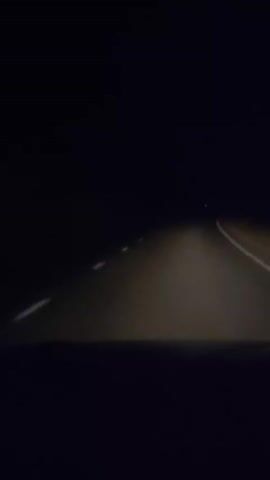 The video shows a first-person view of driving on a dark road at night. The headlights of the vehicle illuminate the asphalt road ahead, with dashed white lines marking the lanes. The surrounding environment is pitch black, indicating a rural or unlit area. There are faint lights visible in the distance, possibly from other vehicles or distant structures. The road appears to be straight for the most part, with a slight curve to the right visible towards the end of the clip. There are no other vehicles or people visible in the immediate vicinity. The footage is grainy due to the low light conditions.