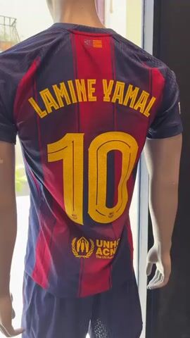 The video displays the back of a mannequin wearing a red and blue striped soccer jersey with yellow lettering. The name "LAMINE YAMAL" is printed across the upper back, and the number "10" is prominently displayed below it. The UNHCR logo is visible on the lower back of the jersey. The mannequin is positioned in front of a wall mural that depicts a soccer stadium filled with cheering fans in yellow and red. A checkout counter with a cash register, card reader, and various items is visible in the foreground, with a red pendant light hanging from the ceiling.