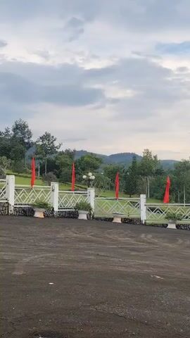 The video shows an outdoor scene with a white fence and red flags in the background. The fence has a patterned design and is decorated with potted plants. Behind the fence, there is a grassy area with trees and hills in the distance. The sky is cloudy, and the lighting suggests it is either early morning or late afternoon. The ground in the foreground is dark and appears to be asphalt or gravel. There are no people visible in the video.