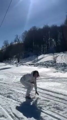 The video shows a woman in a white snowsuit on a sunny day at a snowy location. She is crouching, playing with the snow, and throwing snowballs. There are tracks in the snow suggesting vehicles or snowmobiles have passed through. In the background, there is a line of trees on a snowy hillside.  The sky is blue with the sun shining brightly.
