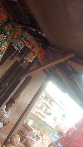The image captures the interior of what appears to be a rustic market stall or small shop, looking out onto an outdoor scene. The ceiling overhead is made of corrugated metal sheets, possibly rusted, supported by exposed wooden beams and planks.

On the upper left side, numerous small, colorful sachets are hanging, likely containing spices, condiments, or snacks. Some of these sachets have visible branding, with "SISA" clearly identifiable on several. The colors are predominantly red, green, and yellow.

Towards the mid-left, a person, possibly a man, is partially visible. He is wearing a light-colored, patterned shirt and appears to be holding or sorting items in a woven basket or container. Below him, in the lower left and central foreground, a stack of round, light-brown vegetables, resembling potatoes or yams, is visible.

A large, open doorway or window on the right side of the frame provides a view of the outside. Beyond the opening, a multi-story building with light-colored walls and blue-tinted windows can be seen in the distance, suggesting a residential or commercial structure. The ground outside appears to be unpaved, possibly dirt, with some scattered objects. In the immediate lower right corner of the frame, a large pile of red material, possibly fabric or plastic, is visible.

The lighting suggests it is daytime, with the interior of the stall being somewhat shadowed, while the view outside is brighter. The interior walls appear to be made of rough, unfinished concrete or plaster.
