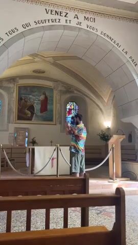 A person wearing a colorful shirt and shorts is standing in a church, holding a camera and filming. The person is facing towards the altar area of the church. The church has pews, an altar, and religious artwork on the walls. There is also text written on the archway above the person, which appears to be in French. Another person is partially visible in the background, near a painting on the wall. The lighting in the church suggests it might be daytime.
