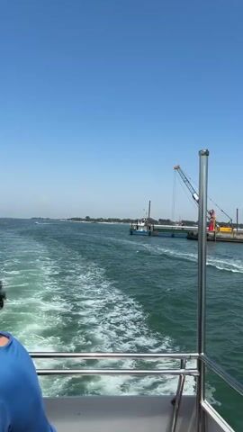 The video shows a view from the stern of a boat moving through a body of water under a clear, blue sky. The water is green, with white wakes from the boat. Metal railings line the stern. A boat with at least four people onboard passes to the right. The boat is mostly blue with a wooden interior. It leaves a white wake as it moves. In the background, a pier or shoreline is visible with a crane and other industrial equipment. The distant land is flat and has some buildings and vegetation.