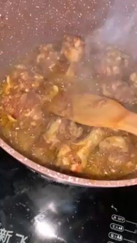 A close-up shot shows a light brown nonstick pan containing chicken pieces and a thick sauce, likely curry. A wooden spoon is used to stir the mixture. Steam rises from the hot food, obscuring the pan's contents. The pan sits on a black induction cooktop.