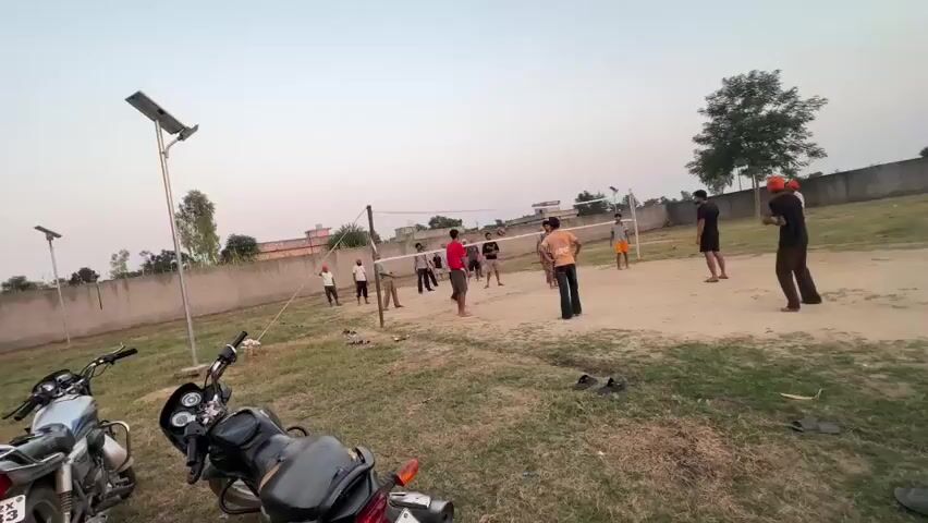 The video shows an outdoor volleyball game. The court is made of dirt and surrounded by grass. There is a net set up for the game, and several people are playing on both sides. The sky is overcast, and the light suggests it is either early morning or late afternoon. In the foreground, there are two motorcycles parked on the grass. In the background, there are some buildings and trees. The people are dressed in casual clothing, and some are wearing turbans. The overall atmosphere appears to be relaxed and social.