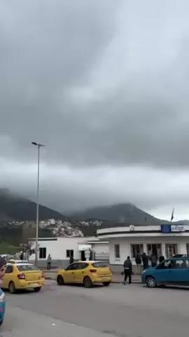 The video shows a street scene with cars parked along the side. There are yellow taxis and a blue car. In the background are mountains shrouded in clouds. A few buildings are visible, including one that appears to be an office or station with people standing near the entrance. An older man in a dark hooded robe walks across the foreground, looking towards the camera. The sky is overcast and grey.