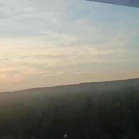 The video displays a time-lapse shot of a sunset viewed from a moving vehicle. The sky transitions from a light blue with wispy clouds to shades of orange and yellow as the sun descends. The horizon is marked by a dark, silhouetted landscape, possibly rolling hills or a forest line. The sun itself appears as a bright orb with a strong lens flare effect extending to the left. The foreground shows a glimpse of a road or highway, with the edge of the vehicle's interior visible at the top of the frame in the initial seconds. There are no people or distinct objects identifiable in the landscape. The overall ambiance is serene and captures the natural phenomenon of sunset.