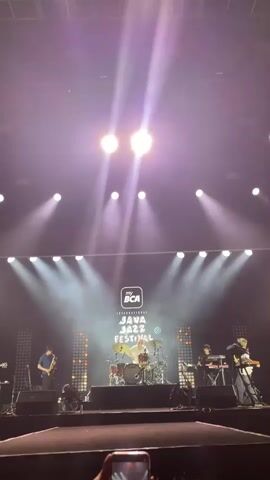 The video shows a band performing on a stage at the "my BCA International Java Jazz Festival". The band consists of a guitarist, a saxophonist, a drummer, and a keyboard player. They are playing under bright stage lights with the festival logo displayed on a screen behind them. An audience member is filming the performance with their phone. The stage is decorated with vertical light panels. The video ends by panning up to show the overhead stage lighting and rigging structure.