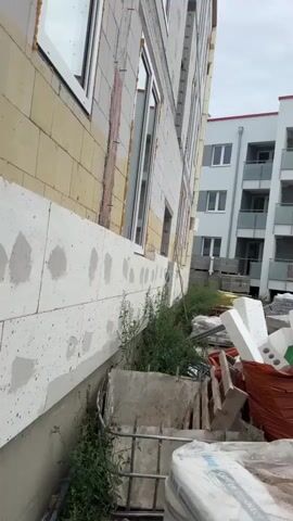 This video shows a construction site. The primary focus is on the exterior of a building under construction, with scaffolding and building materials visible. In the foreground, there are stacks of white insulation blocks and a large orange tarp containing building debris. A concrete cutting saw is also visible. In the background, there is a multi-story residential building with balconies. The overall scene depicts an active construction area with materials and equipment laid out.