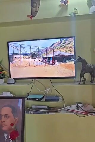 The video shows a flat-screen television displaying a scene of what appears to be an outdoor area with a rustic structure, possibly a shed or small building, surrounded by hills or mountains. There are metal poles supporting a roof-like structure over a section of the ground in front of the building. The area looks like it could be a farm or a rural dwelling. Below the television, on shelves, are various objects, including a framed portrait of a man wearing a suit and tie with a red rose, a small statue of a horse, electronic devices, and other miscellaneous items. Decorative butterflies and a sculpted face are visible on the wall above the television. The overall impression is of a home interior with the television as a focal point, displaying an outdoor scene.