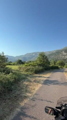 This is a first-person perspective video taken from a motorcycle riding on a paved road. The road is asphalt and appears to be in a rural area. To the left of the road, there is dry grass and some green bushes. Further to the left and in the background, there are more trees and a large mountain range. The sky is clear and blue. To the right of the road, the terrain slopes upwards with vegetation. The motorcycle's handlebar and mirrors are visible in the foreground, suggesting the rider is on a touring motorcycle. The sun appears to be high in the sky, casting shadows. There are no other vehicles or people visible in the immediate vicinity.